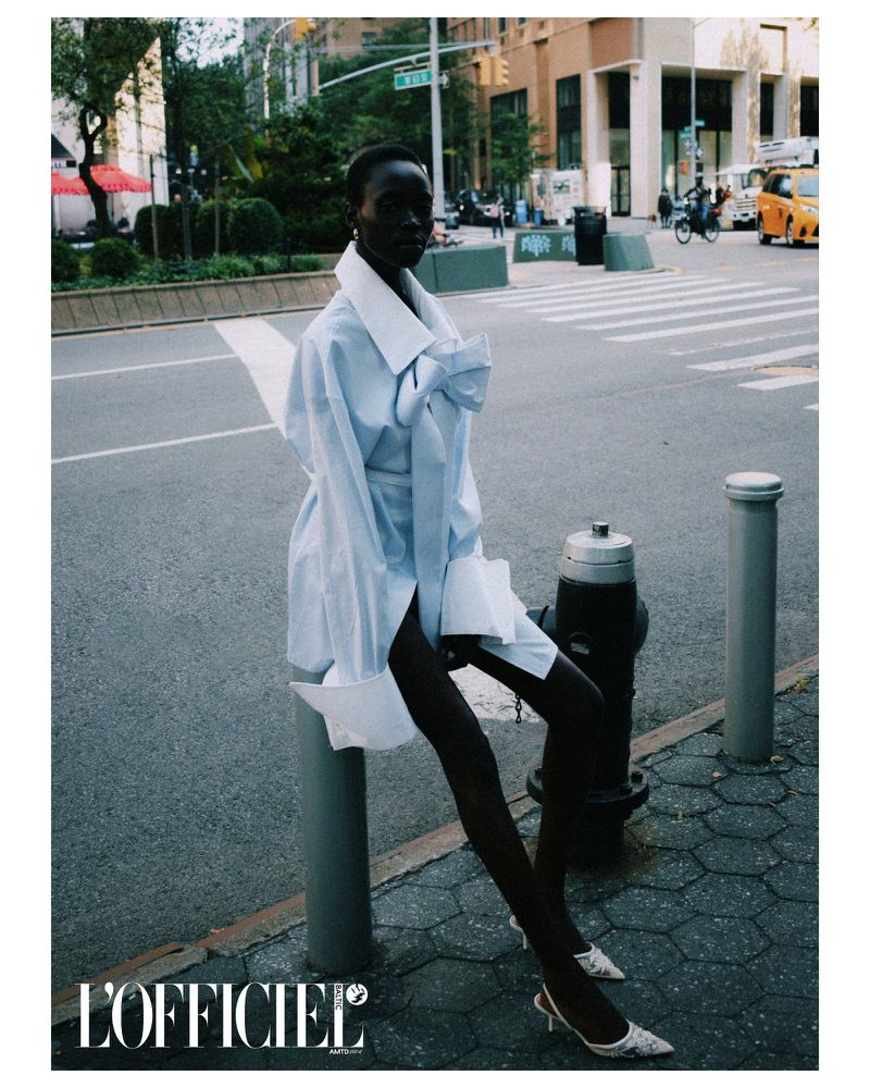 Fashion editorial shot of an African American woman in an oversized blue shirt dress sitting in an urban environment with buildings.