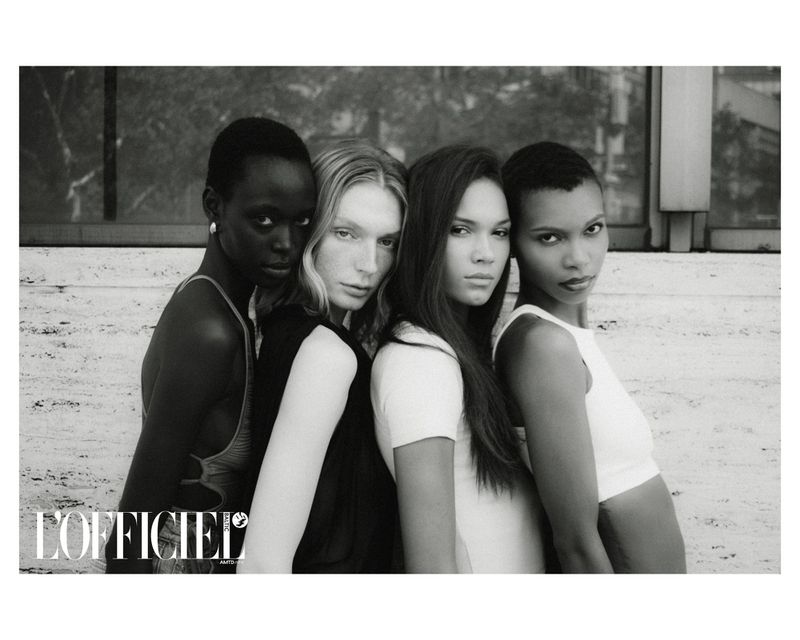 Monochrome group portrait of four diverse women in stylish tops against a textured wall with a window, evoking a high-fashion editorial aesthetic.