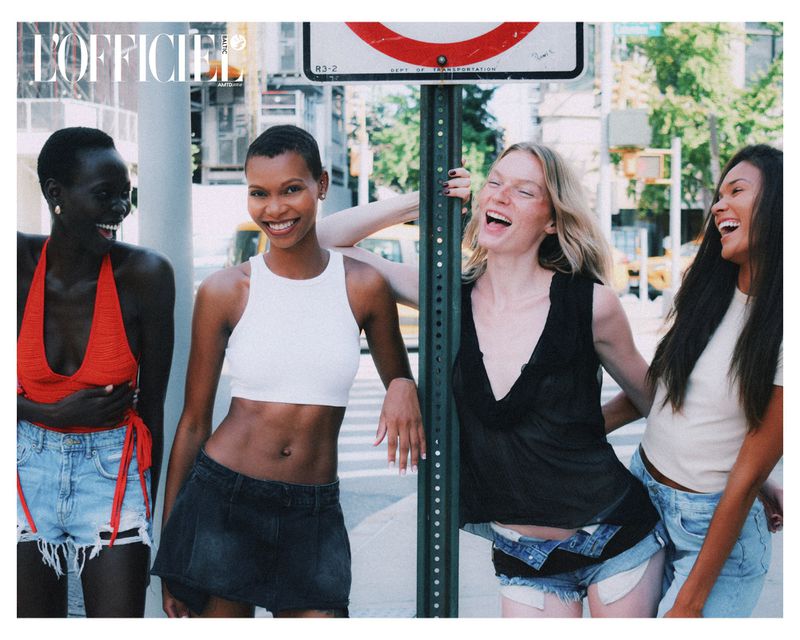 Four diverse women smile and pose in denim shorts and tops on a city street corner near a street sign with bright daylight.