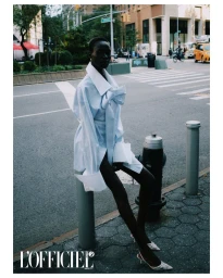 Model wearing an oversized light blue shirt with white cuffs seated on a city street post for an editorial feature.