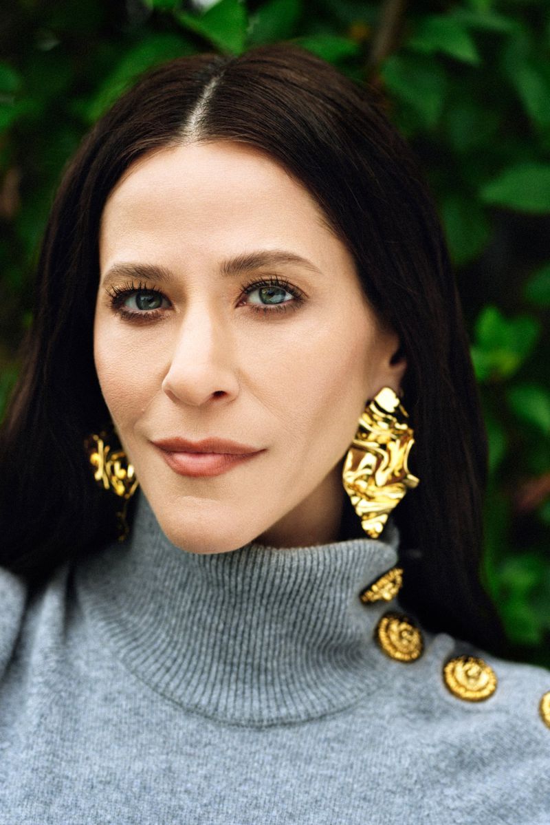 Close-up portrait of a beautiful adult female with dark hair, wearing a gray turtleneck sweater and gold statement earrings in an outdoor setting.