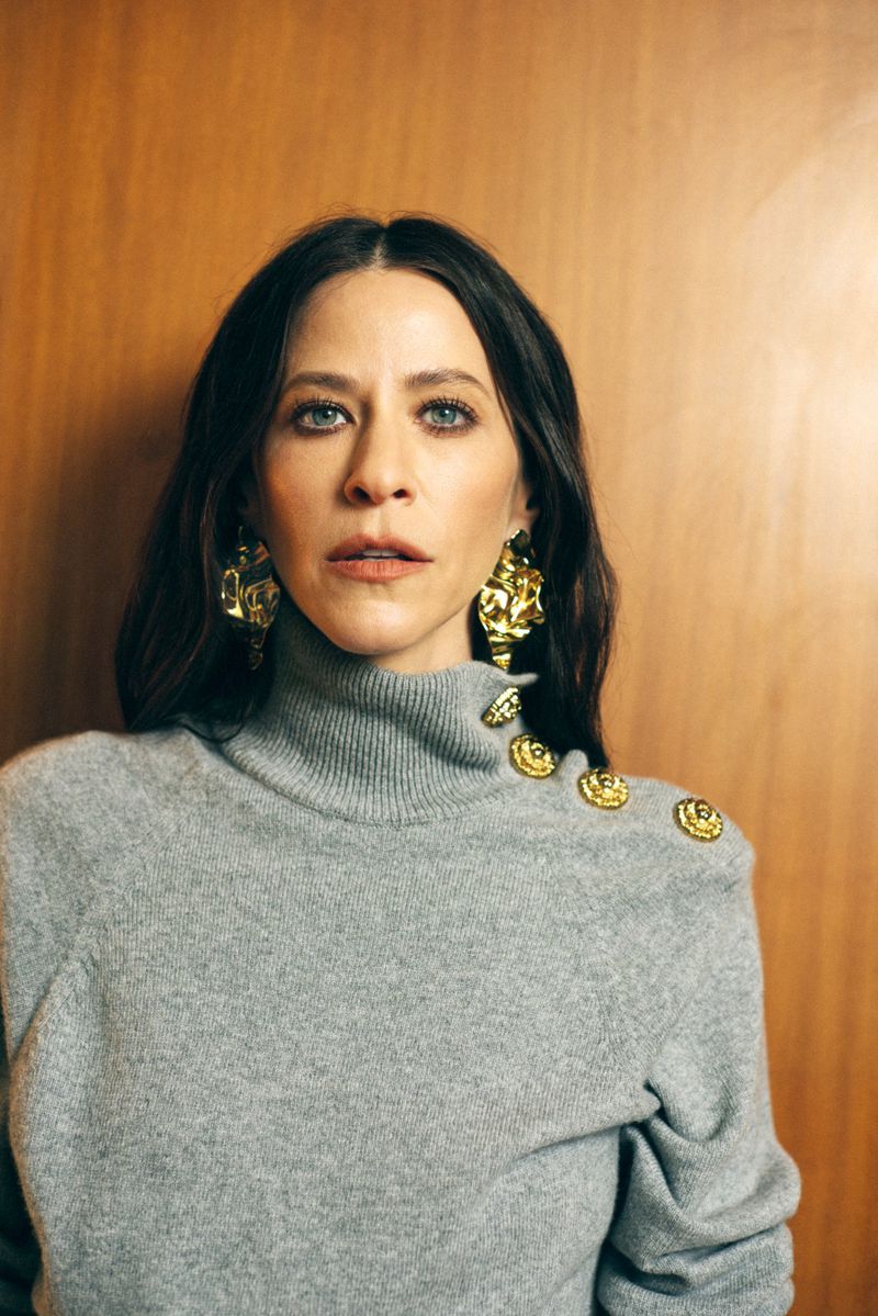 Portrait of a woman with dark hair in a grey turtleneck sweater and large gold earrings against a wood panel background.