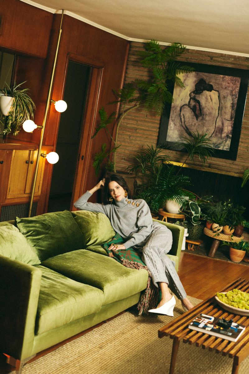 Woman in grey turtleneck and pants relaxes on a green velvet couch in a living room filled with plants and mid-century modern decor.