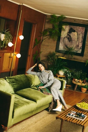 Woman in grey sweater and trousers sitting on a green velvet couch in a retro living room with potted plants.