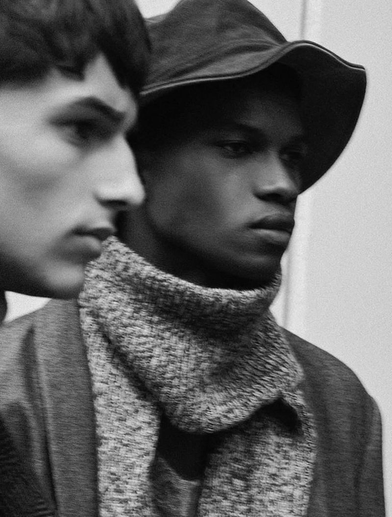 Black and white studio shot of two men in neutral, modern clothing, one wearing a hat and scarf, creating a sophisticated fashion portrait.