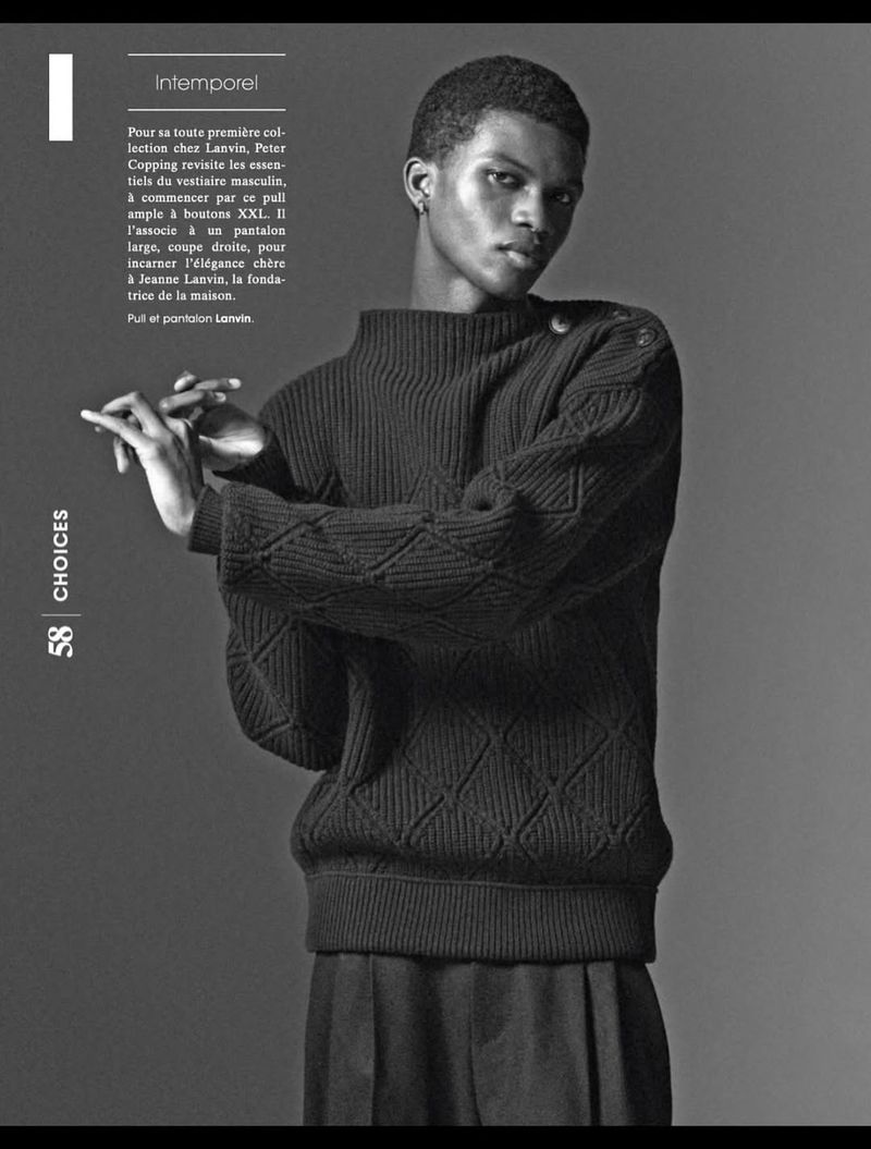 Editorial fashion shot in black and white: a slender Black male model poses in a knit sweater and wide trousers against a grey backdrop.