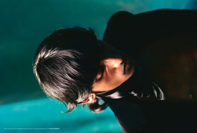 Close-up studio shot of an adult with dark hair and light skin, wearing a black top, against a teal backdrop with moody lighting.