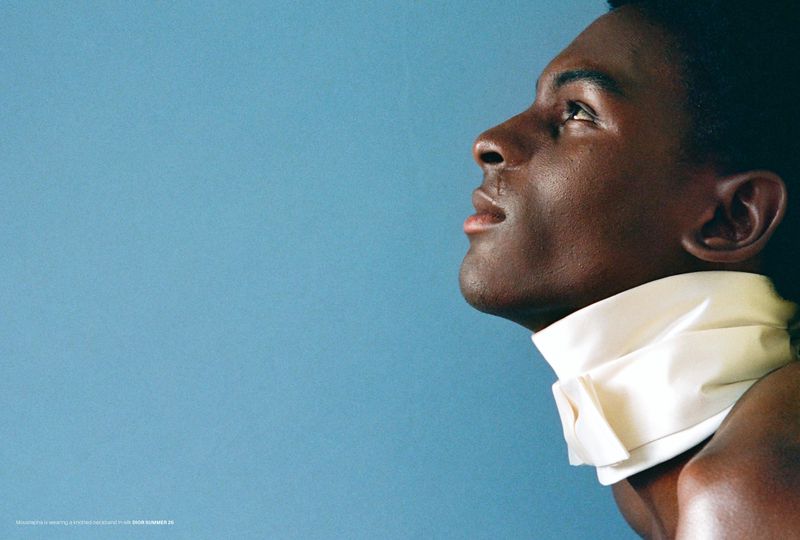 Portrait of an African American male model in profile wearing a white high-necked garment against a light blue studio backdrop.  