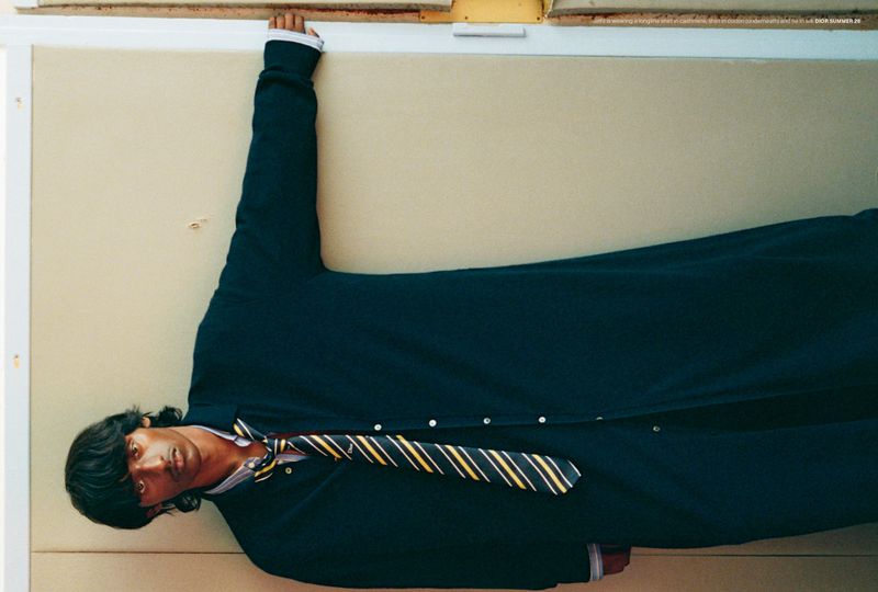 Young man wearing school uniform with striped tie lies on his side. The background is a cream wall with white trim and tan accents.