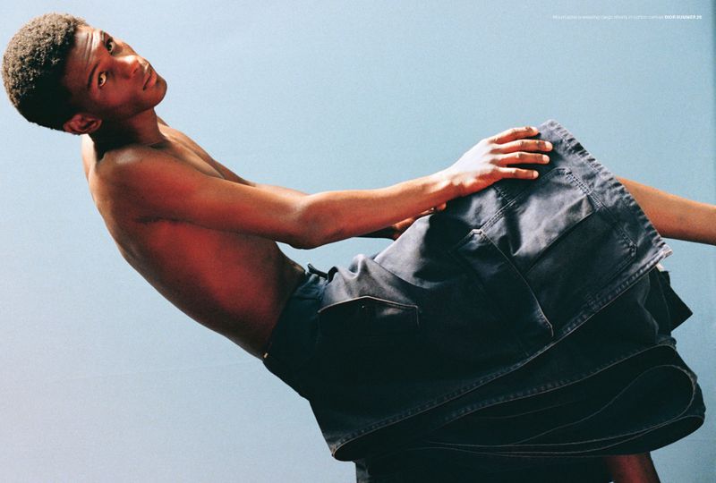 Shirtless male model poses at an angle in denim skirt with a light blue backdrop in a fashion editorial studio shot.