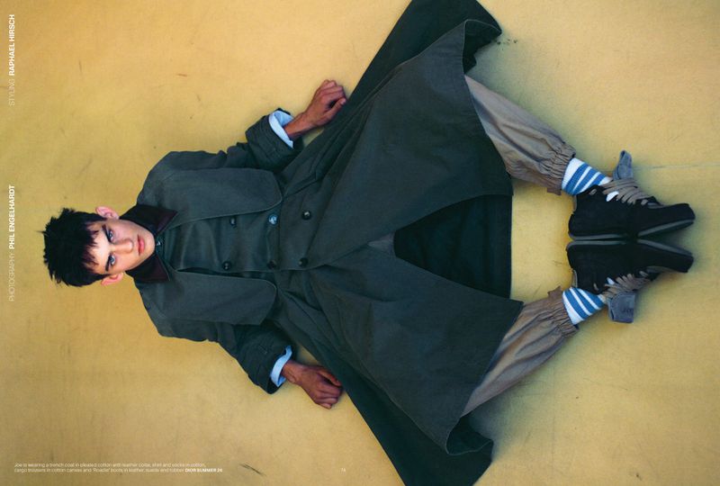 Fashion shot of a young adult man in a dark green trench coat, striped socks, and sneakers, lying on a yellow surface and looking at the camera.