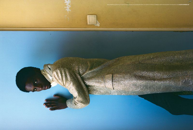 A Black man with dark hair and dark eyes lies on a light blue surface wearing a gold jacket, posing in an indoor environment with yellow wall.