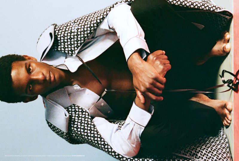 Close-up of a dark-skinned man in a white dress shirt and patterned jacket, lying down and holding his feet, against a light blue backdrop.