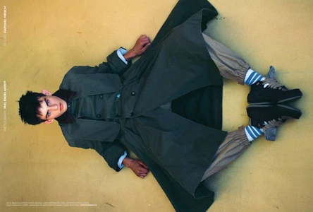 A young male model lying on a yellow surface wearing a dark trench coat, tan cargo trousers, blue and white striped socks, and sneakers.