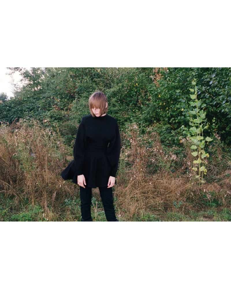 Model Thea Almqvist standing outdoors in an all-black long-sleeve tunic and trousers against overgrown greenery.