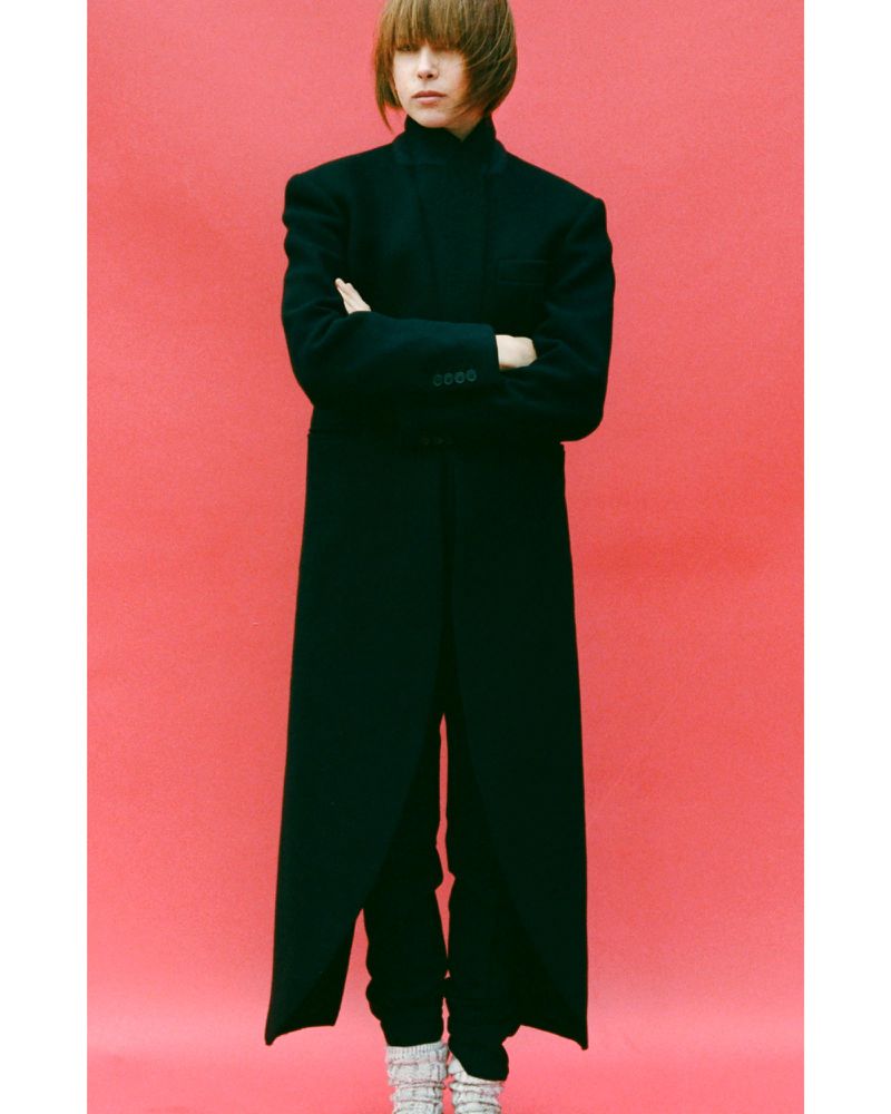 Model with short brown hair wearing a floor-length black coat and white boots against a vibrant pink seamless background.
