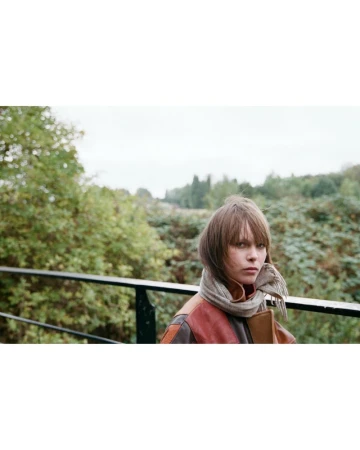 Woman with bangs wearing a scarf and leather jacket in a forest setting, captured in a medium shot with soft, natural lighting.