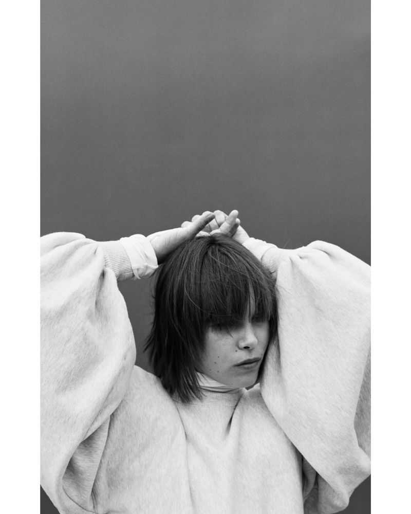 Monochrome portrait of a woman with hands over her head, wearing a high-neck top with puffy sleeves against a gray backdrop.