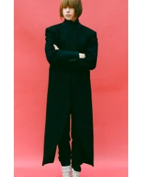 Model with short brown hair wearing a floor-length black coat and white boots against a vibrant pink seamless background.