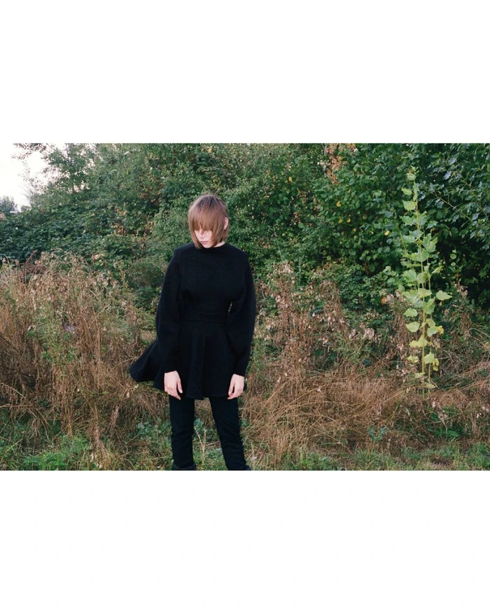Model Thea Almqvist standing outdoors in an all-black long-sleeve tunic and trousers against overgrown greenery.