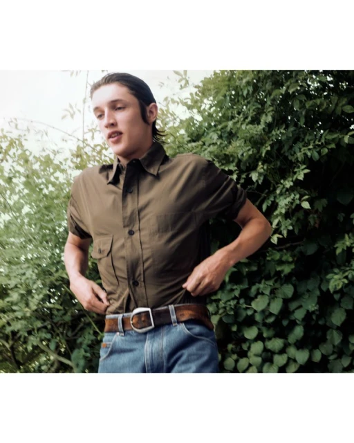 Young man in casual attire adjusts his shirt outdoors surrounded by lush green foliage, captured in a medium shot with natural lighting.