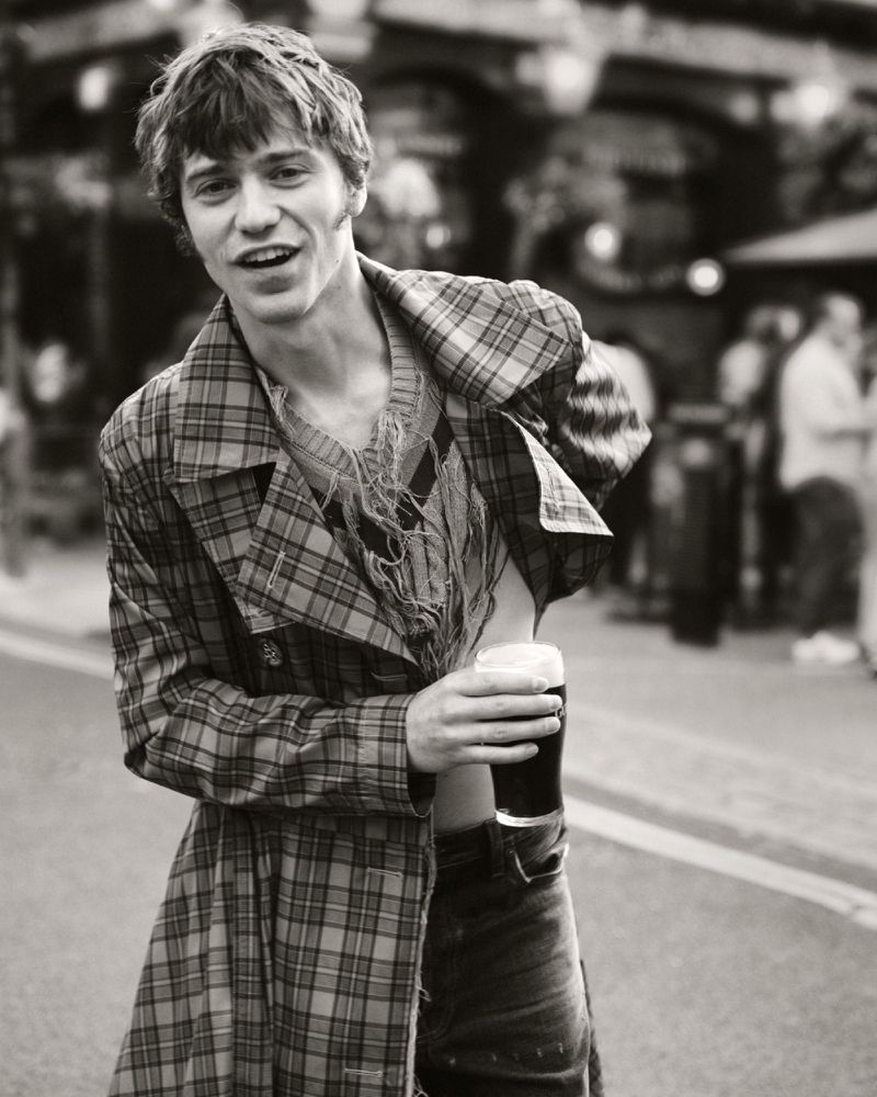 Monochrome portrait of a young adult male smiling, wearing a plaid coat and jeans, holding a dark beer in an outdoor setting.