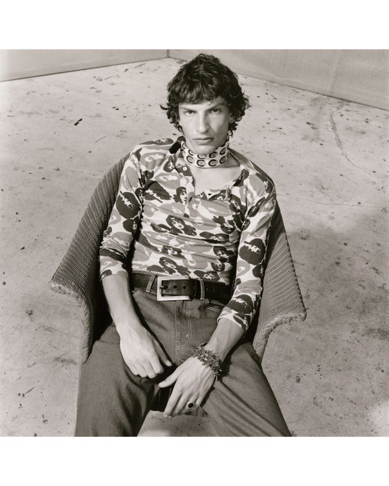 Monochrome portrait of a slender man in a patterned shirt seated in a woven chair against a simple wall background, showcasing a modern aesthetic.