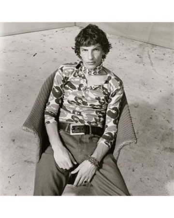 Monochrome portrait of a slender man in a patterned shirt seated in a woven chair against a simple wall background, showcasing a modern aesthetic.