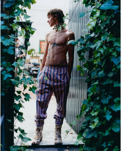 Shirtless adult male in patterned trousers and boots poses near ivy, looking left, with a person in the background indoors.