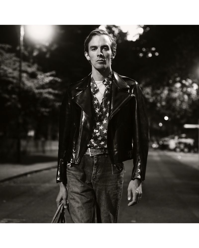 Black and white portrait of a man wearing a leather jacket and star-patterned shirt walking down a street at night with streetlights.