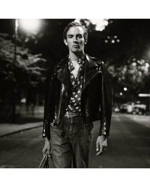 Black and white portrait of a man wearing a leather jacket and star-patterned shirt walking down a street at night with streetlights.