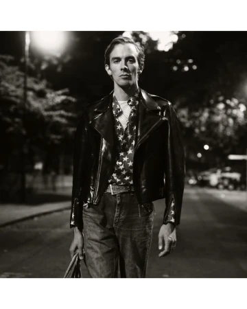 Black and white portrait of a man wearing a leather jacket and star-patterned shirt walking down a street at night with streetlights.