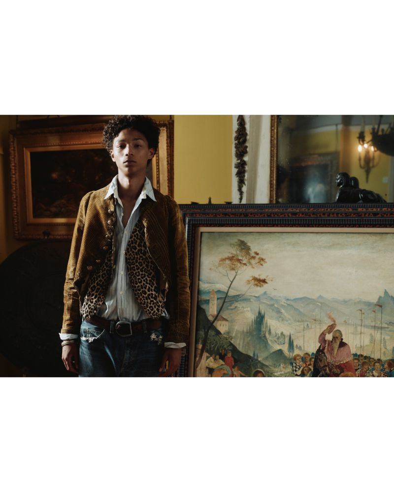 Adult male poses indoors wearing a patterned shirt, jeans, and brown jacket next to an antique landscape painting in a brown frame.
