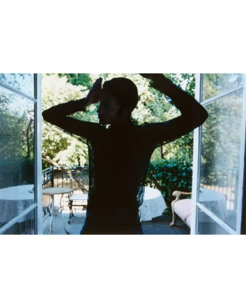 Silhouette of a male model posing in front of open double doors leading to a sunlit balcony with tables and lush greenery.