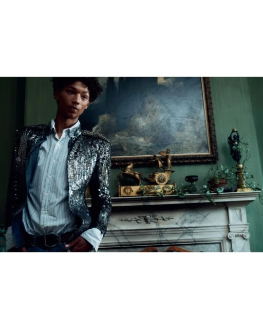 Person in sequined jacket poses near marble fireplace mantel with antique clock, framed painting, and decorative candlesticks.