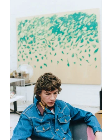 Young man in denim jacket sitting in chair with green and beige art on wall, creating a relaxed fashion portrait in a studio setting.