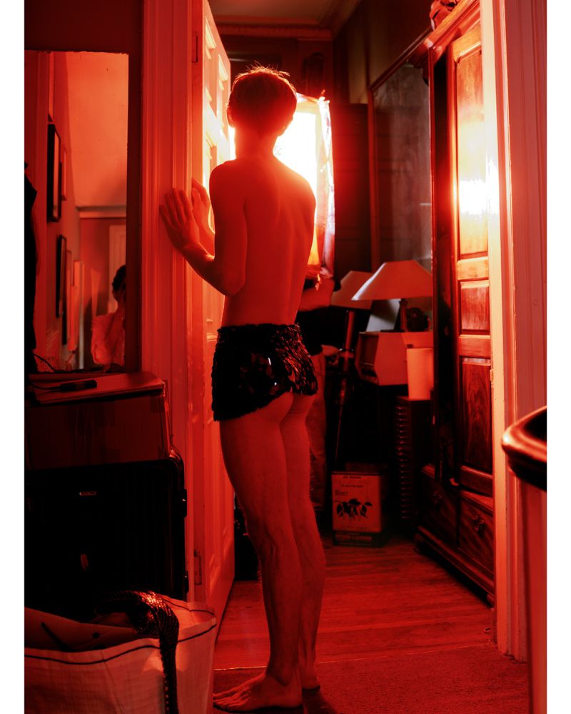 Backlit shirtless person in short skirt stands in red-lit hallway. Wardrobe, doorway, mirror, and bag visible in background.