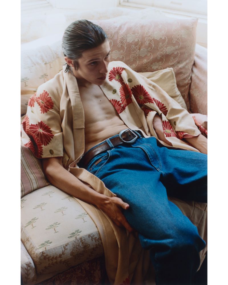 Man with unbuttoned floral robe and blue jeans sits on a patterned couch in an indoor setting with soft, diffused natural lighting.