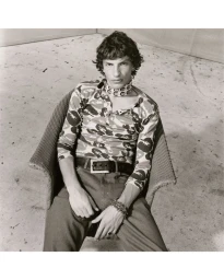 Monochrome portrait of a male model sitting in a woven armchair wearing a printed long-sleeve top, choker scarf, and belted pants.