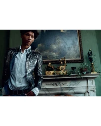 A young man wearing a sequined jacket over a white striped shirt stands before a marble fireplace and dark painting.