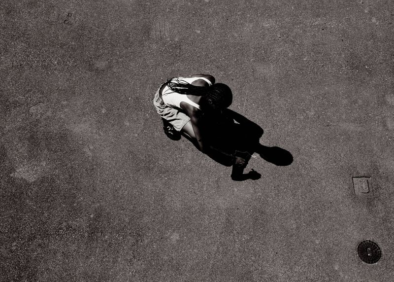 High-angle, black and white shot of a person in shorts and a tank top on a cracked concrete surface, casting a long shadow.
