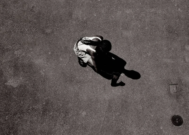 High-angle, black and white shot of a person in shorts and a tank top on a cracked concrete surface, casting a long shadow.