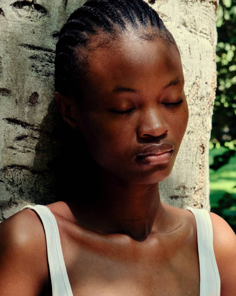 Close-up of an African American woman with braided hair standing next to a tree with her eyes closed, wearing a white tank top.