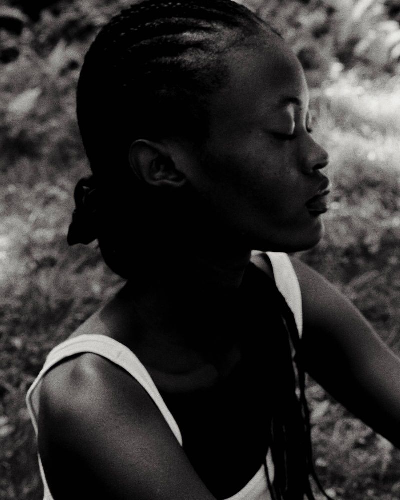 Monochrome portrait of a young Black woman with braided hair, eyes closed, wearing a tank top, set against a blurry, natural background.