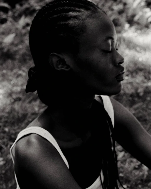 Monochrome portrait of a young Black woman with braided hair, eyes closed, wearing a tank top, set against a blurry, natural background.