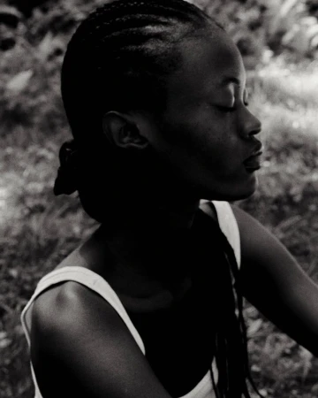 Monochrome portrait of a young Black woman with braided hair, eyes closed, wearing a tank top, set against a blurry, natural background.