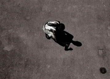 High-angle black and white shot of a person crouching on an asphalt surface, casting a long dark shadow.
