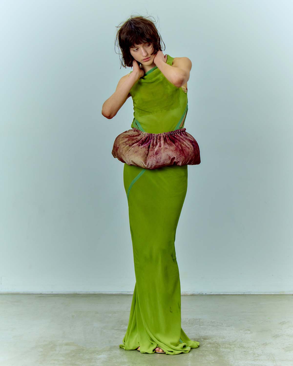 Full-length shot of a woman wearing a green gown with a voluminous pink and brown waist detail, posing against a light blue background.