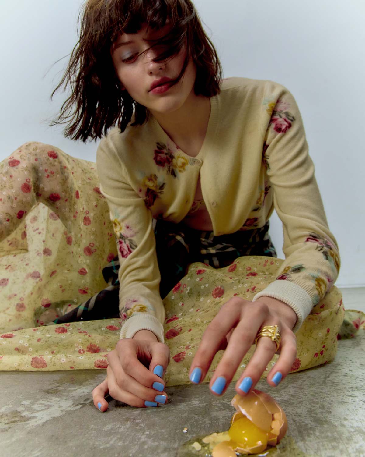 Young woman with bob haircut and blue nail polish poses near a broken egg in a fashion editorial. Floral-patterned shirt and cardigan.