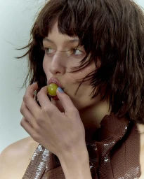Close-up portrait of a woman with dark wavy hair eating a green grape wearing a brown sequined top